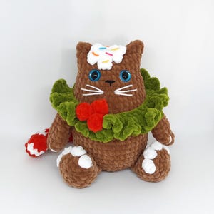 May include: A brown crocheted cat plushie with blue eyes, white whiskers, and a green wreath with red berries. The cat has white frosting with sprinkles on its head and white accents on its paws. It holds a red and white candy cane.