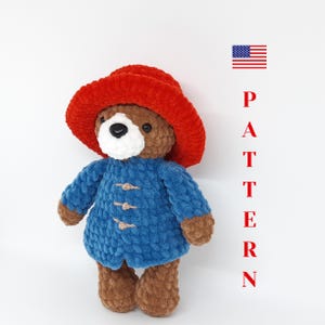 Könnte beinhalten: Ein handgehäkelter Bär mit rotem Hut und blauem Mantel mit Holzknebeln. Der Bär hat eine weiße Schnauze und braunes Fell. Das Wort "PATTERN" steht vertikal in roten Buchstaben neben dem Bären. Der Bär ist etwa 25 cm groß.