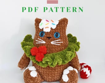 May include: A brown crocheted cat plushie with blue eyes, white whiskers, and a green ruffle collar adorned with red berries. The cat has a white frosting and sprinkle detail on its head. The text "PDF PATTERN" is above the cat.