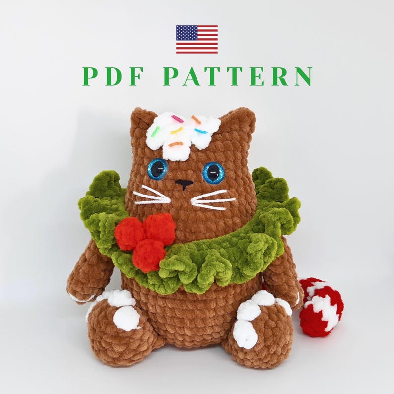 May include: A brown crocheted cat plushie with blue eyes, white whiskers, and a green ruffle collar adorned with red berries. The cat has a white frosting and sprinkle detail on its head. The text "PDF PATTERN" is above the cat.