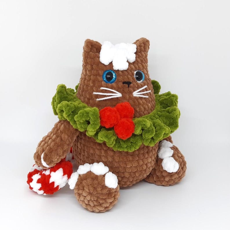 May include: A brown crocheted cat plushie with blue eyes, white whiskers, and a white patch on its head. It wears a green wreath with red berries and holds a red and white candy cane. The cat is a festive holiday decoration.