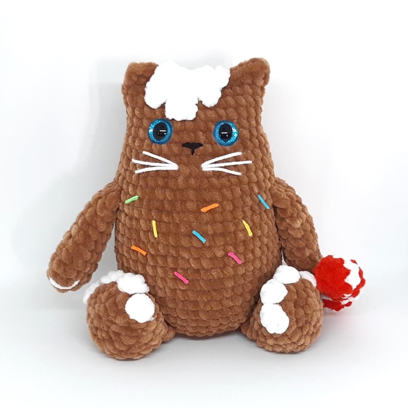 May include: A handmade brown crochet cat plushie with blue eyes, white whiskers, and a white frosting-like design on its head. The plushie has colorful sprinkles and holds a red and white ball. The cat is sitting.