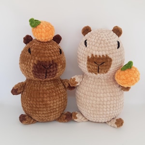 May include: Two crocheted plush capybara toys, one brown with an orange crocheted fruit on its head, and one beige holding an orange crocheted fruit.