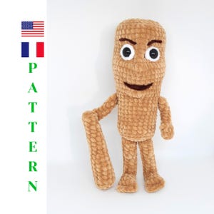 May include: A tan, crocheted plush figure with a textured, yarn-based surface. The figure has large, round black and white eyes, dark brown eyebrows, and a wide, dark brown mouth. It features long arms and legs, with one arm extended downward.