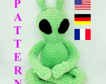 May include: Green crocheted alien plush toy sitting with legs crossed. The toy has black eyes and a textured surface. The word "PATTERN" is written in pink letters on the left side of the image. The flags of the United States, Germany, and France are on the right side of the image.