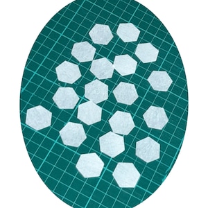 May include: A close-up of a green cutting mat with white hexagon-shaped fabric pieces scattered on top.