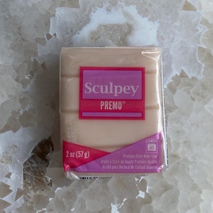 May include: A 2 ounce (57 gram) block of Sculpey Premo oven-bake clay. The clay is a light beige colour and is packaged in a clear plastic wrapper with pink and white labelling. The label includes the brand name, product name, weight, and instructions for use.