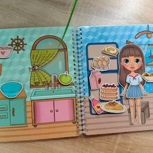 Pode incluir: Um livro de atividades infantil com espiral, com tema de cozinha. As ilustrações incluem uma boneca, eletrodomésticos de cozinha, alimentos e uma cena de janela. A paleta de cores apresenta pastéis e cores brilhantes, criando um ambiente lúdico.