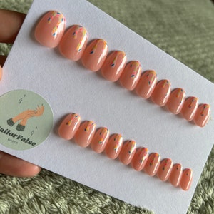 May include: A set of 10 nude-coloured false nails with a white and colourful sprinkle design. The nails are oval shaped and have a glossy finish.