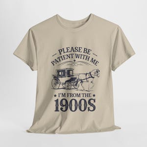 Op de afbeelding: Beige T-shirt met een zwart-witte illustratie van een koets getrokken door een paard. De tekst "Please be patient with me I'm from the 1900s" staat op het shirt gedrukt.