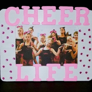 May include: White picture frame with pink letters spelling "CHEER LIFE" and pink rhinestones around the border. A photo of a group of young girls in cheerleading uniforms is inside the frame.