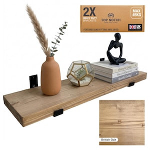 A wooden floating shelf in British Oak, supported by black brackets, displaying a terracotta vase with dried plants, a geometric terrarium, and books. The shelf includes "2X Heavy Duty Brackets" and "Max 45kg" text. Fixtures and fittings included.