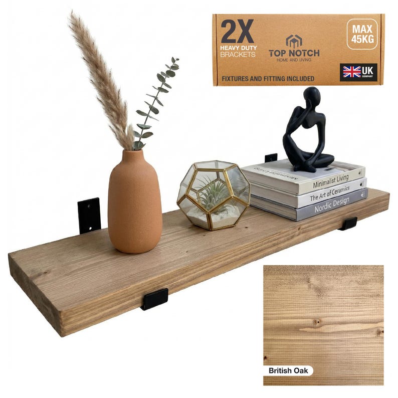 A wooden floating shelf in British Oak, supported by black brackets, displaying a terracotta vase with dried plants, a geometric terrarium, and books. The shelf includes "2X Heavy Duty Brackets" and "Max 45kg" text. Fixtures and fittings included.