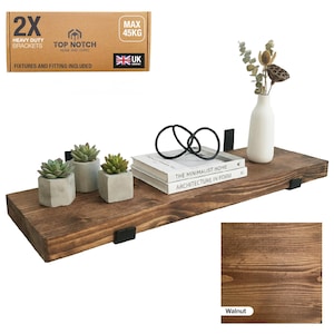 A walnut-stained wooden floating shelf with black metal brackets. The shelf holds small potted succulents, books, and black decorative rings. The box in the image says "Heavy Duty Brackets" and "Max 45KG".