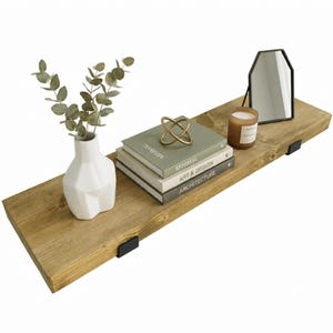 May include: A rectangular wooden floating shelf with two black metal brackets. On the shelf sits a white geometric vase with eucalyptus, a stack of three books titled Minimal, Art & Design, and Architecture, a gold wire sphere, a candle, and a mirror.