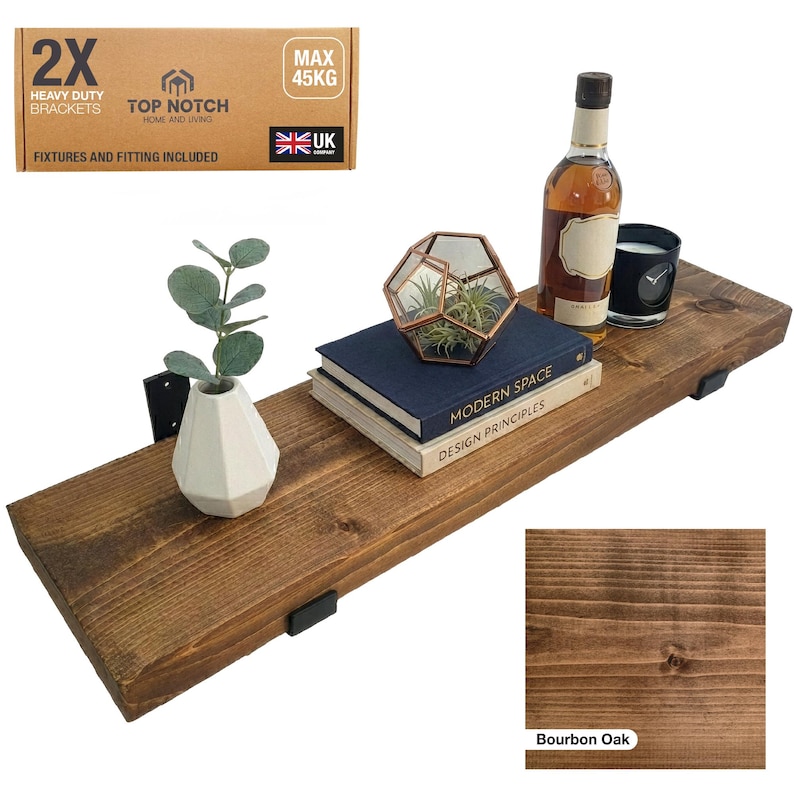 A Bourbon Oak Stain dark brown wooden shelf with black brackets, holding decorative items like a geometric terrarium, books, a candle, and a bottle. The shelf is made of Bourbon Oak. The image also shows a box with the text "2X HEAVY DUTY BRACKETS".