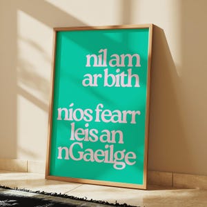 Könnte beinhalten: Ein gerahmter Druck mit einem türkisfarbenen Hintergrund und weißem Text in fetter Schrift. Der Text lautet "nil am ar bith nios fearr leis an nGaeilge".