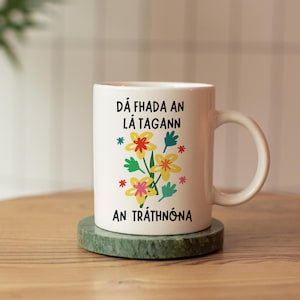 以下が含まれることがあります： 白いセラミック製のマグカップに、花柄のデザインと「Dá fhada an lá tagann an tráthnóna」というテキストが書かれています。