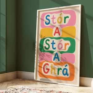 Puede incluir: Un cuadro rectangular con marco que presenta el texto Stór A Stór A Ghrá en letras coloridas, redondeadas y estilo burbuja. Las palabras están dispuestas verticalmente sobre un fondo crema, separadas por formas orgánicas onduladas en rosa, amarillo, verde y naranja.