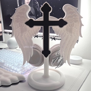May include: A white headphone stand featuring a black cross design with attached angel wings. The stand is on a white base, with a pair of headphones hanging from it. The text "17BLK Studio" is visible.