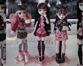 Könnte beinhalten: Drei Modepuppen mit blasser Haut und rosa-schwarzem Haar, die jeweils unterschiedliche Outfits tragen. Die linke Puppe trägt ein Barett und einen gemusterten Rock, die mittlere ein rosa Rüschenoberteil und die rechte hält eine kleine Fledermausfigur. Alle stehen auf rosa, spinnennetzförmigen Sockeln.