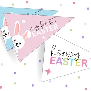 May include: Three pastel-coloured triangle pennants with Easter greetings. The top pennant is light blue with the text "My First Easter". The middle pennant is pink with the text "My First Easter" and a white bunny peeking out from behind the text. The bottom pennant is white with the text "Hoppy Easter" in rainbow colours.
