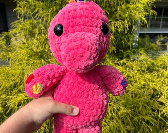 May include: A handmade pink crochet dinosaur plushie with large black eyes and a colorful horn. The dinosaur's feet and arms have colorful accents. The plushie is held in front of a green bush.