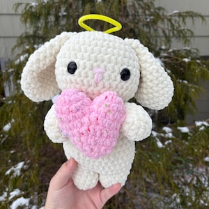 May include: A white crocheted bunny with a pink heart and a yellow halo. The bunny is holding the heart in its paws.