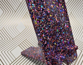 May include: A phone stand made of resin with a glittery finish. The stand is a dark purple color with red, blue, and gold glitter embedded in the resin. The stand is shaped like a triangle with a flat base and a slanted top.