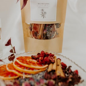 May include: A brown paper bag with a clear window showing dried orange slices, cinnamon sticks, lavender, and rose petals. The bag has a white label that says "ALL MY LOVE" and "tea witch collection" with a floral design.