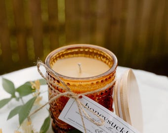 Puede incluir: Vela de vidrio color ámbar con tapa de madera, atada con hilo de bramante, y una etiqueta que dice "honeysuckle collective". La vela está sobre una superficie blanca con hojas verdes y flores amarillas.