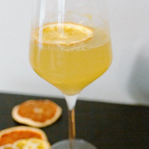 May include: A glass of yellow citrus drink with a dried orange slice garnish. The glass is on a black surface with dried orange slices around it.