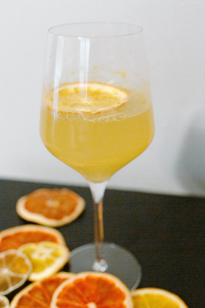 May include: A glass of yellow citrus drink with a dried orange slice garnish. The glass is on a black surface with dried orange slices around it.
