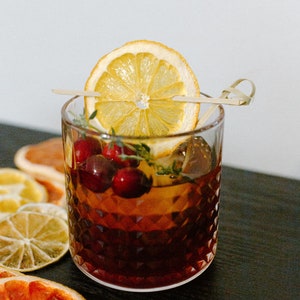 May include: A close-up of a cocktail in a rocks glass with a dried orange slice garnish and cranberries. The drink is dark brown in colour and has a layer of ice. The glass is sitting on a black surface with dried citrus slices.