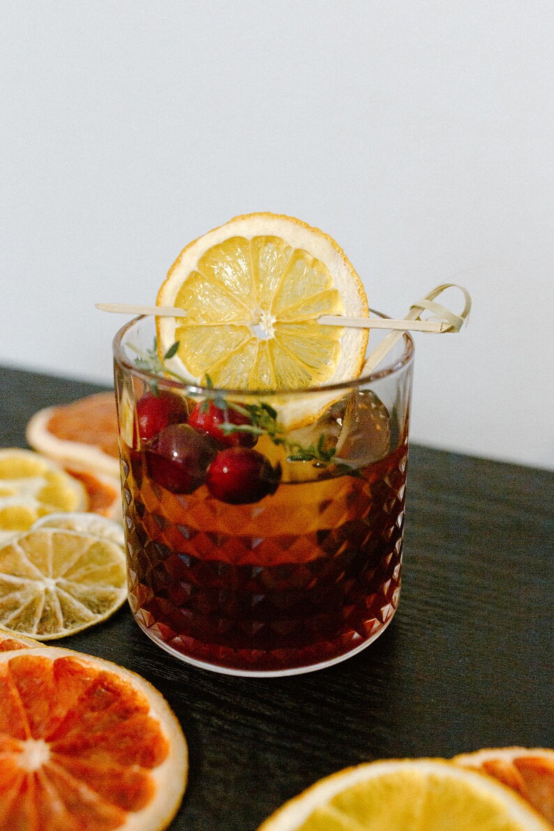 May include: A close-up of a cocktail in a rocks glass with a dried orange slice garnish and cranberries. The drink is dark brown in colour and has a layer of ice. The glass is sitting on a black surface with dried citrus slices.