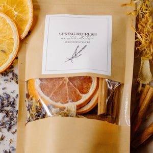 May include: A brown paper bag with a clear window, filled with dried orange slices, cinnamon sticks, and lavender. The bag has a label that reads "Spring Refresh" and "sea witch collective floral simmer pot kit."
