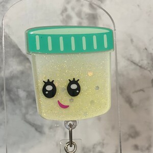 Pode incluir: Um porta-crachá amarelo claro e brilhante com um design de recipiente de desenho animado com um rosto sorridente e uma tampa turquesa. O porta-crachá tem uma parte traseira de plástico transparente e um clipe de metal prateado.