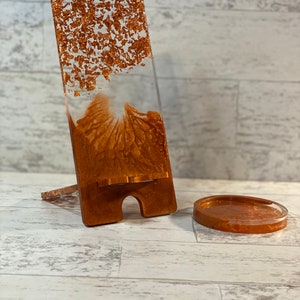 May include: A copper-colored resin phone stand with a matching coaster. The stand is rectangular with a slot for a phone and a small support leg. The resin has a clear base with copper leaf accents.