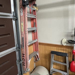 Puede incluir: Una escalera roja y plateada de Louisville está apoyada contra una pared. La escalera es una escalera de mano con varios peldaños. Un pequeño taburete plateado y otras herramientas están en el suelo.