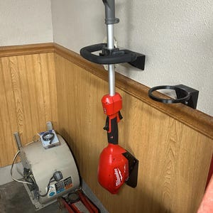 May include: A red and black Milwaukee string trimmer mounted on a wall bracket. The trimmer is hanging vertically, with a black handle and a red battery pack. A second bracket holds a black circular object. The wall is wood-paneled.