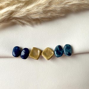 May include: A collection of six handmade ceramic beads in a row. The beads alternate between a deep navy blue and a metallic gold color. The shapes include oval and square designs. The background is a white surface.