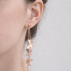 May include: Rose gold butterfly earrings. These dangle earrings feature a small ball stud and delicate chain with three butterfly charms. The earrings are shown against a neutral background.