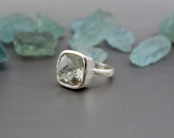 May include: A silver ring with a large, square-cut, pale green gemstone. The ring has a textured band and setting, and the gemstone is faceted. The background features light blue, irregularly shaped glass pieces.