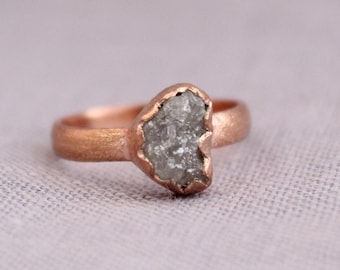 May include: A rose gold ring featuring a rough-cut, gray-colored diamond. The ring's band has a smooth, matte finish, and the diamond is set in a textured bezel. The overall design is organic and unique, suitable for everyday wear or special occasions.