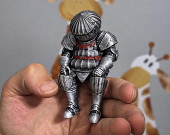 May include: A small, detailed figurine of a knight in full silver-colored plate armor, sitting in a slumped posture. The armor features a rounded helmet, segmented shoulder pauldrons, and a red belt strap across the chest and waist.