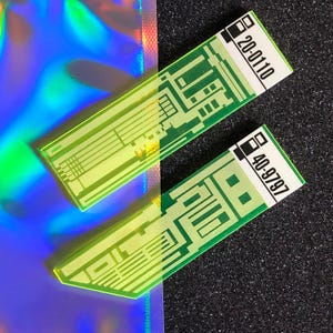 May include: Two rectangular, translucent green acrylic pieces with circuit board designs. Each has a white label with black text, one reading "20-0110" and the other "40-9797". The background includes a holographic iridescent surface and a black textured surface.