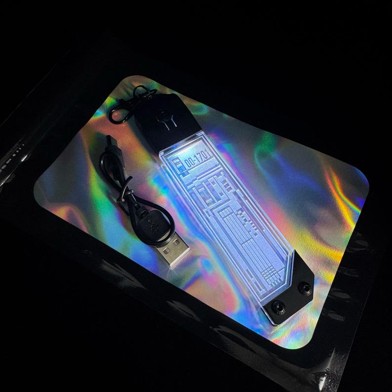 K&ouml;nnte beinhalten: Ein leuchtendes, rechteckiges Ger&auml;t mit dem Text "00-1701" auf einer holografischen Oberfl&auml;che. Das Ger&auml;t hat eine schwarze Ober- und Unterseite mit einem USB-Kabel. Das Ger&auml;t emittiert blaues und wei&szlig;es Licht.