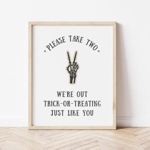May include: A framed sign with the text "PLEASE TAKE TWO" and "WE'RE OUT TRICK-OR-TREATING JUST LIKE YOU." The sign features a black and beige skeleton hand making a peace sign.