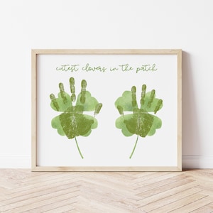 Puede incluir: Impresión enmarcada con dos tréboles verdes hechos con huellas de manos sobre un fondo blanco. El texto "cutest clovers in the patch" está escrito en verde sobre los tréboles. El marco es de madera clara.