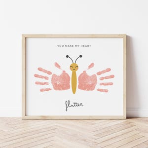 Peut inclure: Une impression encadrée avec un papillon rose fait d'empreintes de mains. Le papillon a un corps jaune et un visage souriant. Le texte "You make my heart flutter" est imprimé sous le papillon.